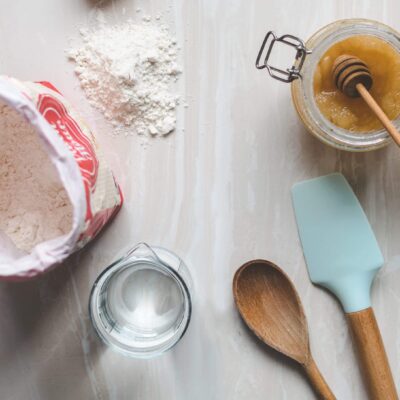A table with flour, honey, water, a plastic spatula and wooden spoon on it