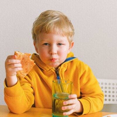 Boy Wearing Yellow Hoodie Eating Pizza And Holding A Glass Of Water